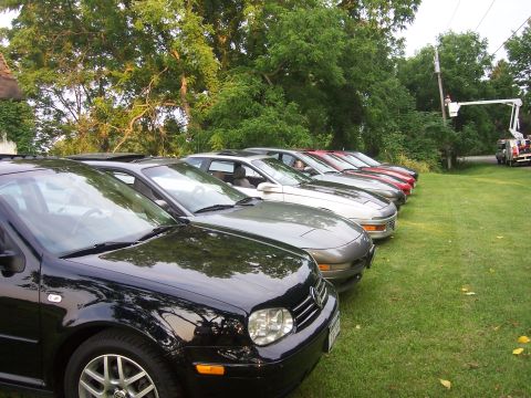 1996 Ford Probe - Multiple vehicles are parked in a line on a lawn with trees and a utility truck in the distance.