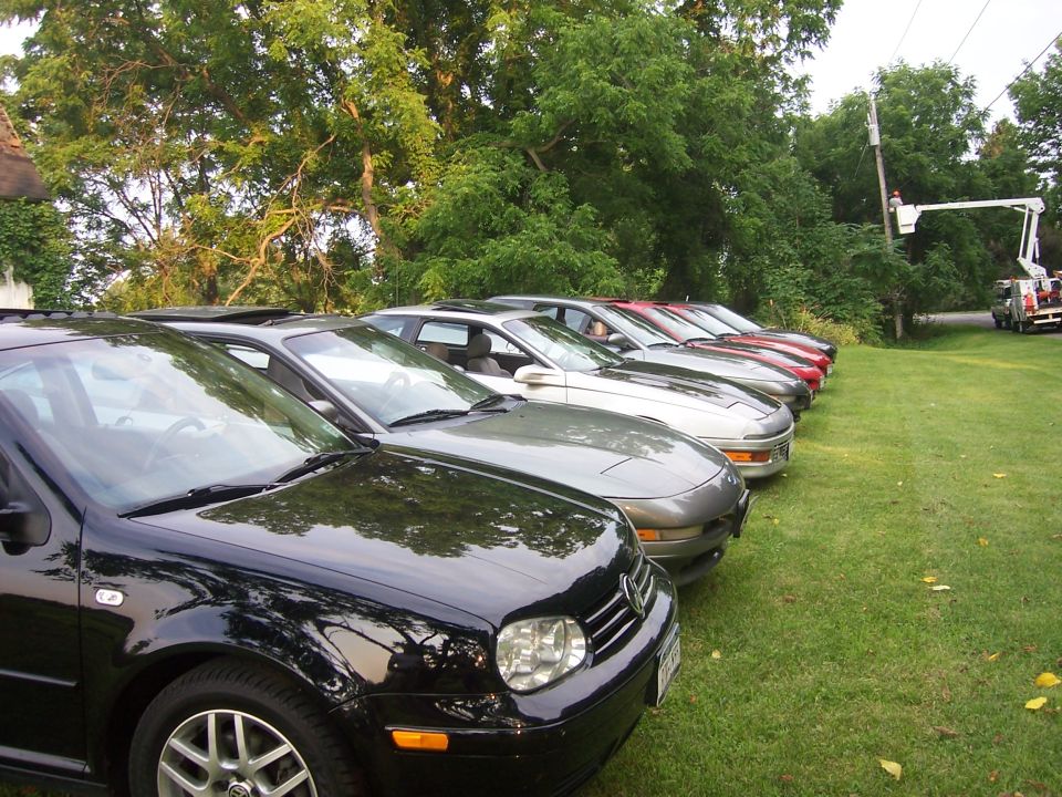Several cars parked in a row on a grassy area with trees in the background