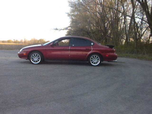 Dark red Ford sedan in profile on pavement, rear spoiler, chrome multi-spoke wheels, tinted windows