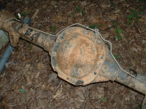 1985 Ford Bronco - A rusty removed rear axle housing lies on leaf-covered ground, photographed from above showing the bolt-on differential cover, spring perch…