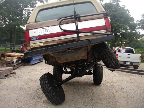 1987 Ford Bronco - Rear view of a lifted 1987 Ford Bronco on a trailer, with a white pickup truck behind it.