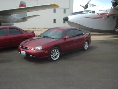 1998 Ford Taurus - A red 1998 Ford Taurus is parked next to a large airplane, with another red car visible behind it.