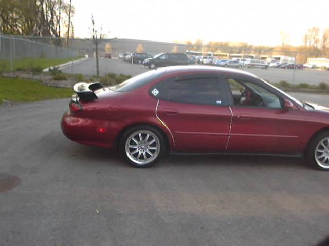 Right side view of a dark red Ford Taurus sedan with rear spoiler and aftermarket alloy wheels in a parking lot