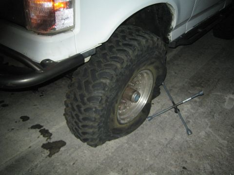 1990 Ford Bronco - mud-terrain tire on a chrome wheel of a white lifted truck sits beside a four-way lug wrench on pavement at night.