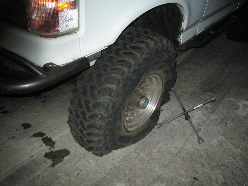 Front wheel of a white lifted truck at night with a knobby off-road tire and a four-way lug wrench on the ground