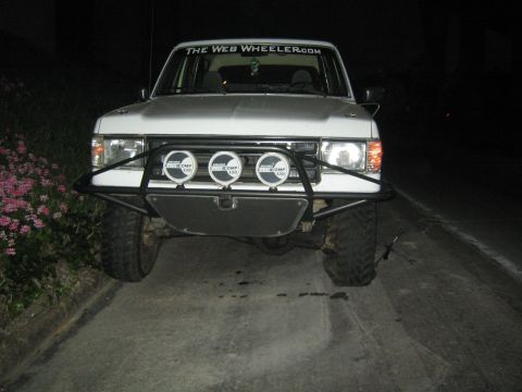 1990 Ford Bronco - silver lifted truck photographed head-on at night with a tubular front bumper, three round driving lights, and a TheWebWheeler.com windshie…