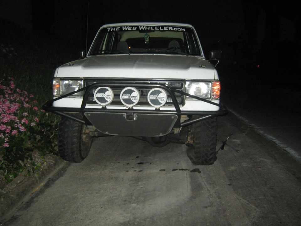 Front of a silver lifted truck at night with a tube bumper and three round driving lights, windshield banner reads TheWebWheeler.com