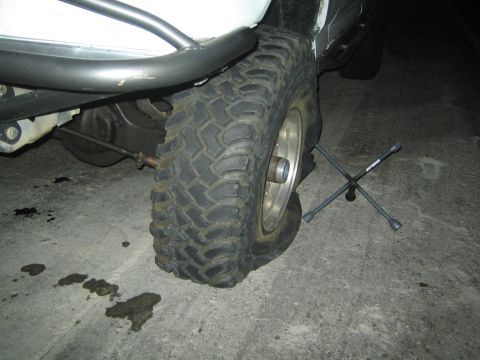 1990 Ford Bronco - Close-up of a lifted truck's front mud-terrain tire and exposed suspension at night, with a four-way lug wrench lying on the pavement.