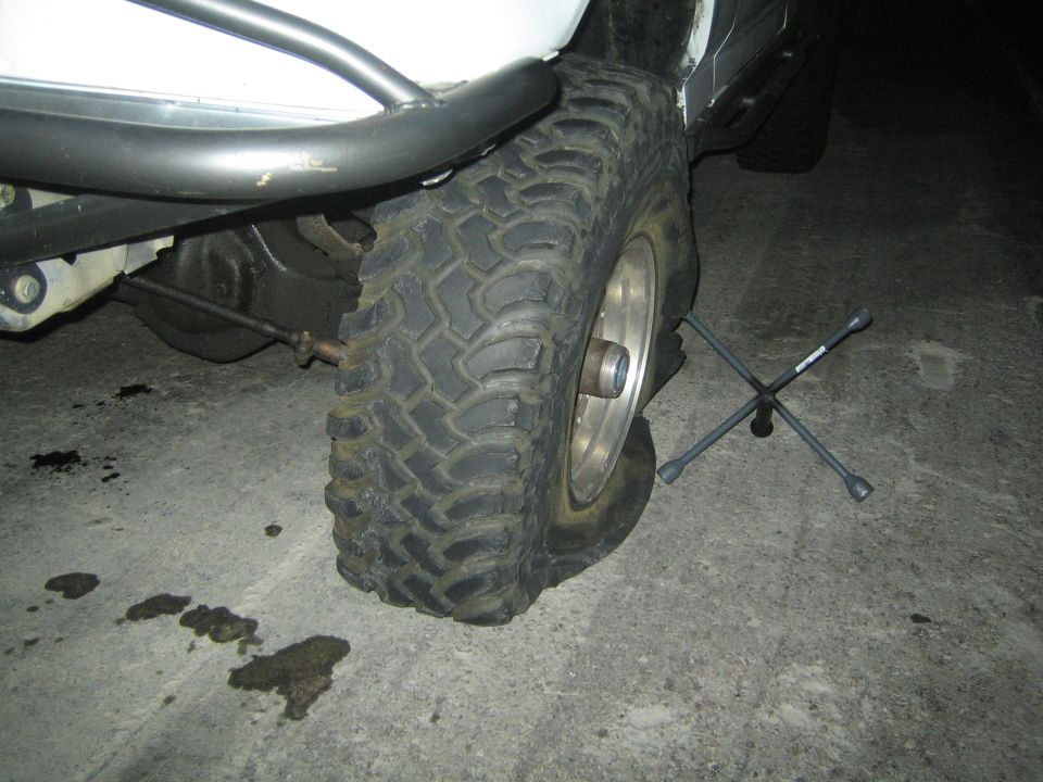 Front wheel and suspension of a lifted truck at night showing a mud tire, chrome wheel and a four-way lug wrench on the ground