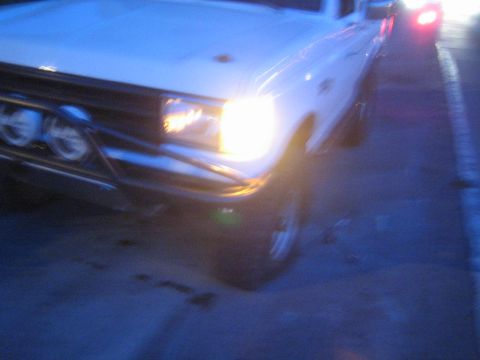1990 Ford Bronco - motion-blurred dusk shot of a white truck's front corner, headlight glowing, with a tubular bumper holding round driving lights.