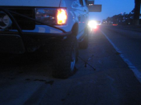 1990 Ford Bronco - white lifted truck sits on a highway shoulder at dusk, parking light glowing, with a four-way lug wrench on the pavement and traffic approa…