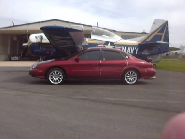 Red Ford Taurus sedan side view parked in front of a blue and white Navy airplane at an airport