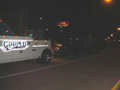 1992 Ford Bronco - A tow truck is parked on the side of the road at night, with a dark SUV behind it.