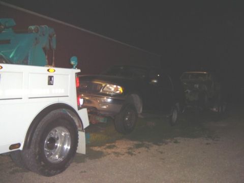 1992 Ford Bronco - A tow truck is preparing to haul away a dark pickup truck at night, with another vehicle visible behind it.