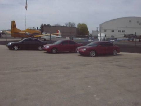 1998 Ford Taurus - Three sedans are parked in front of an airplane and industrial buildings under an overcast sky.