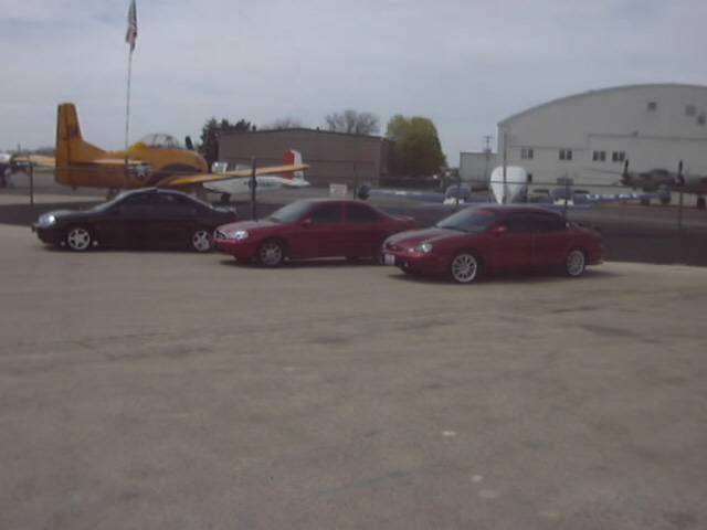 Three red and black sedans parked outdoors with an airplane and buildings in the background