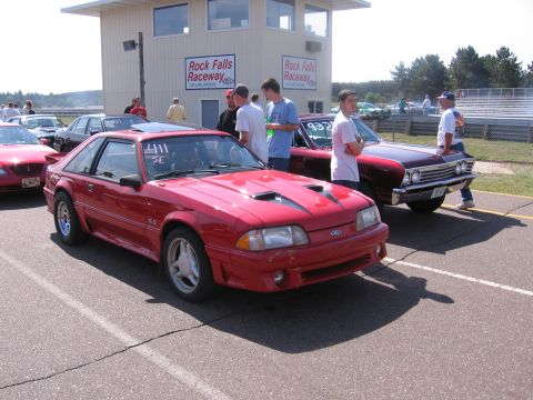 1989 Ford Mustang - A red 1989 Ford Mustang is parked at a raceway, with other vehicles and spectators present.