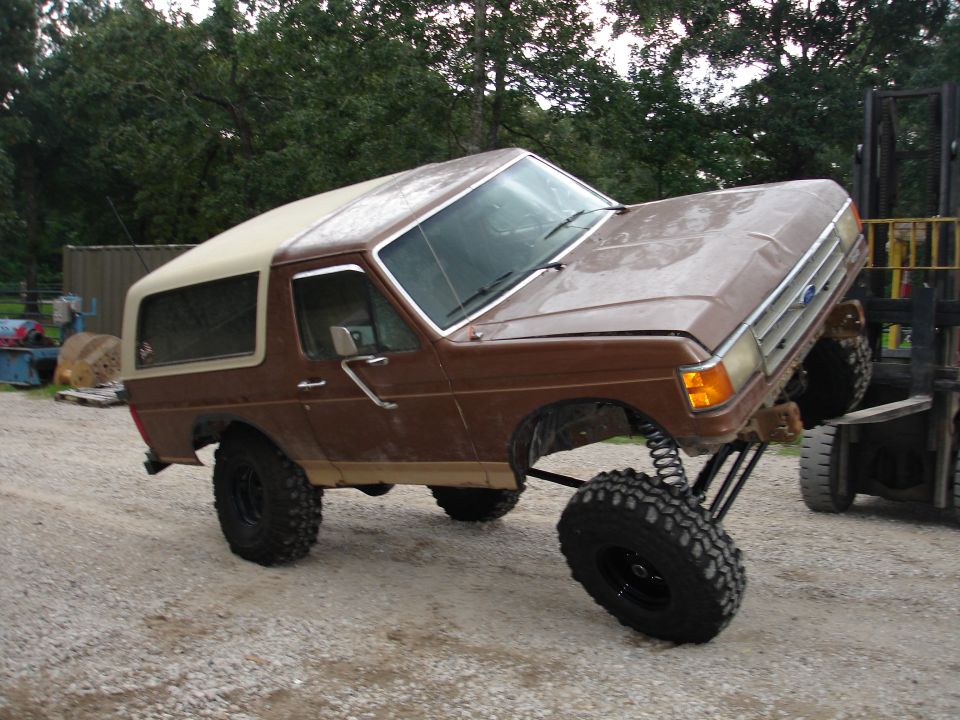 Lifted brown Ford Bronco with tan top flexing on big mud tires beside a forklift in a gravel lot