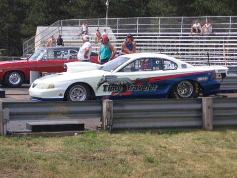 1989 Ford Mustang - Two drag cars are lined up at a race track, with spectators watching from the stands.