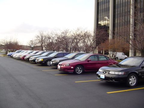 1998 Ford Taurus - Several sedans are parked in a lot next to a modern office building.