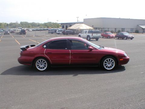 1998 Ford Taurus - A red 1998 Ford Taurus sedan is parked in a lot with other vehicles and buildings in the background.