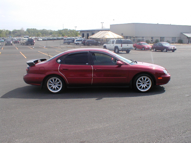 Red 1998 Ford Taurus sedan with a rear spoiler and aftermarket wheels, viewed from the side