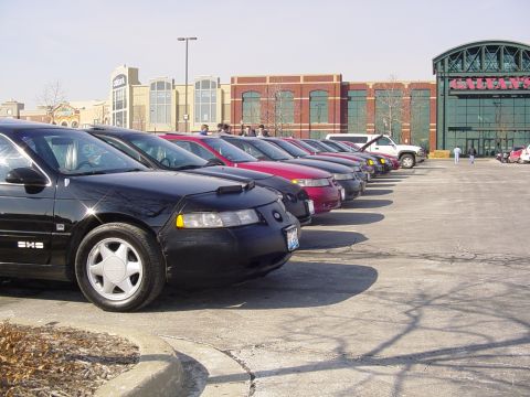 1998 Ford Taurus - A line of sedans, including a black 1998 Ford Taurus, are parked in a lot outside a shopping center.