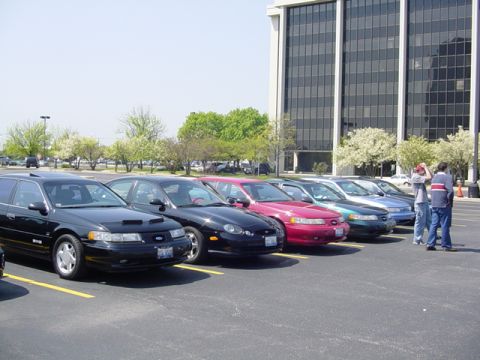 1998 Ford Taurus - A row of Ford Taurus sedans, including a 1998 model, are parked outside a modern office building.