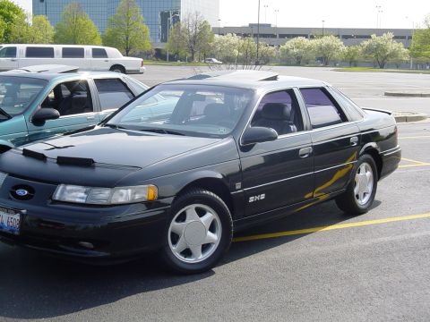 1998 Ford Taurus - A black 1998 Ford Taurus is parked next to a teal sedan, with a limousine and office buildings in the background.