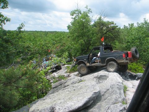 1992 Ford Bronco - Two off-road vehicles tackle a challenging rocky path surrounded by dense green foliage.