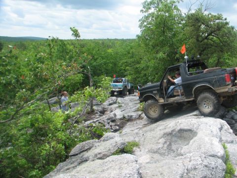1992 Ford Bronco - Off-road vehicles navigate a challenging rocky path through a dense, green forest under a cloudy sky.