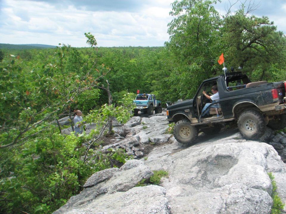 Two off-road vehicles driving over large rocks on a wooded trail. The lead vehicle is black with an orange flag