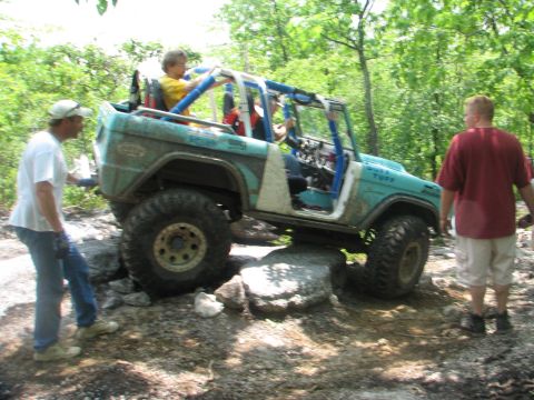 1992 Ford Bronco - A teal off-road vehicle with a roll cage is shown navigating a rocky trail with people inside and around it.