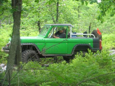 1992 Ford Bronco - A green off-road vehicle navigates a wooded trail, its driver visible behind the wheel.