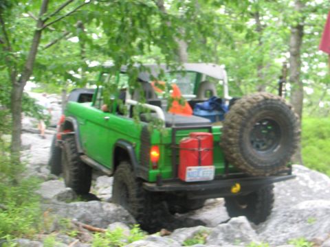 1992 Ford Bronco - Green off-road SUV navigates rocky terrain with a spare tire and red gas can mounted on the rear.