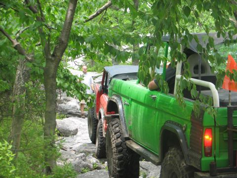 1992 Ford Bronco - Two off-road vehicles navigate a challenging rocky trail surrounded by lush green trees.