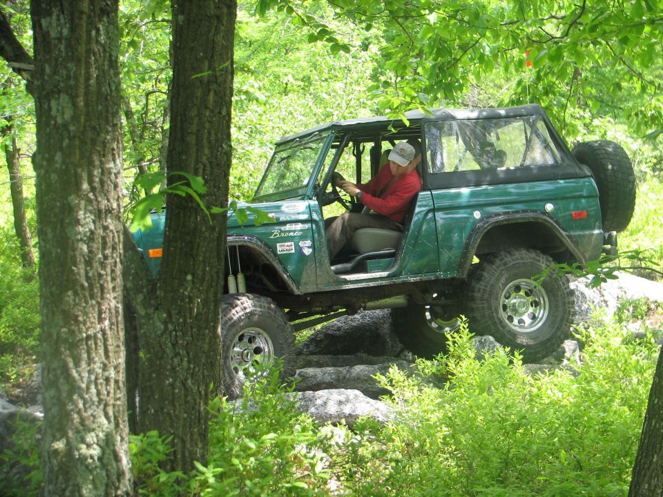 Green Ford Bronco driving over rocks on a wooded trail. Driver wears a red shirt and grey hat