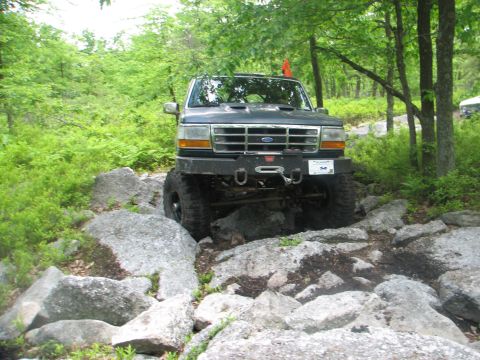 1992 Ford Bronco - This Ford Bronco is shown off-roading on a challenging rocky path, surrounded by lush green foliage.