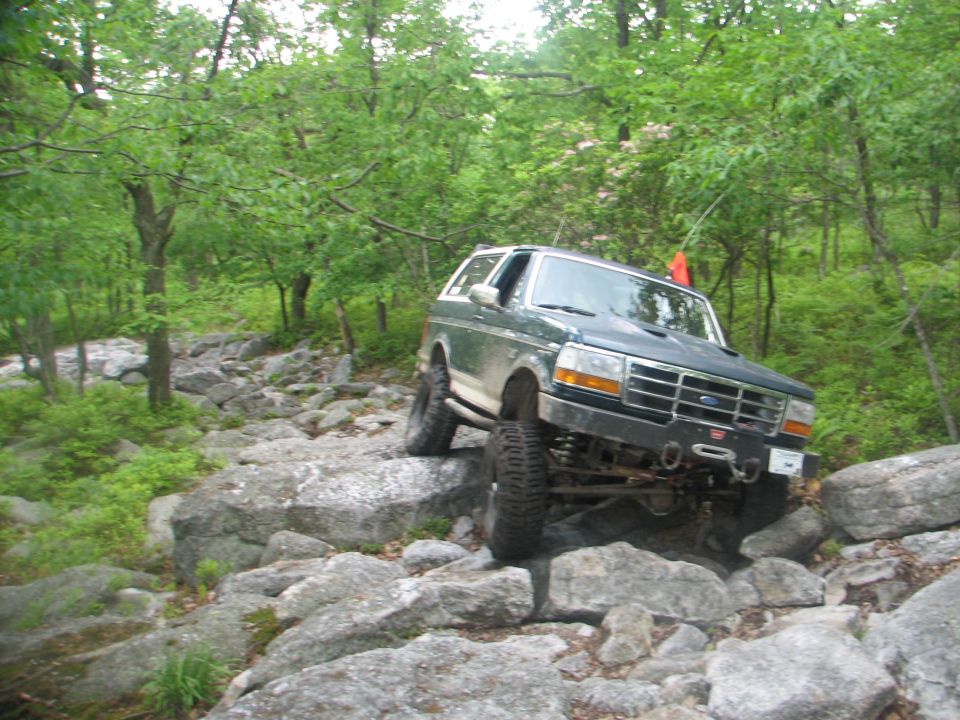 Green Ford Bronco flexing over boulders on a wooded trail with front tire lifted