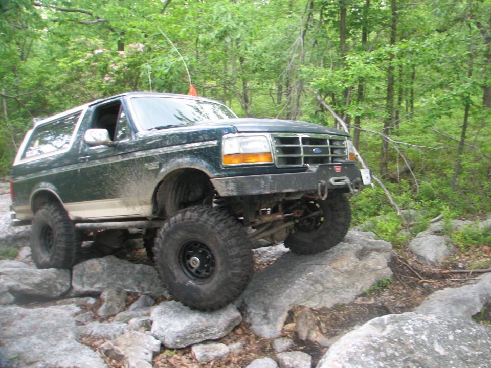 Lifted green Ford Bronco on 35s crawling over boulders on a wooded rock trail