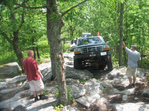1992 Ford Bronco - A group of off-road vehicles tackle a challenging rocky terrain in a wooded area.