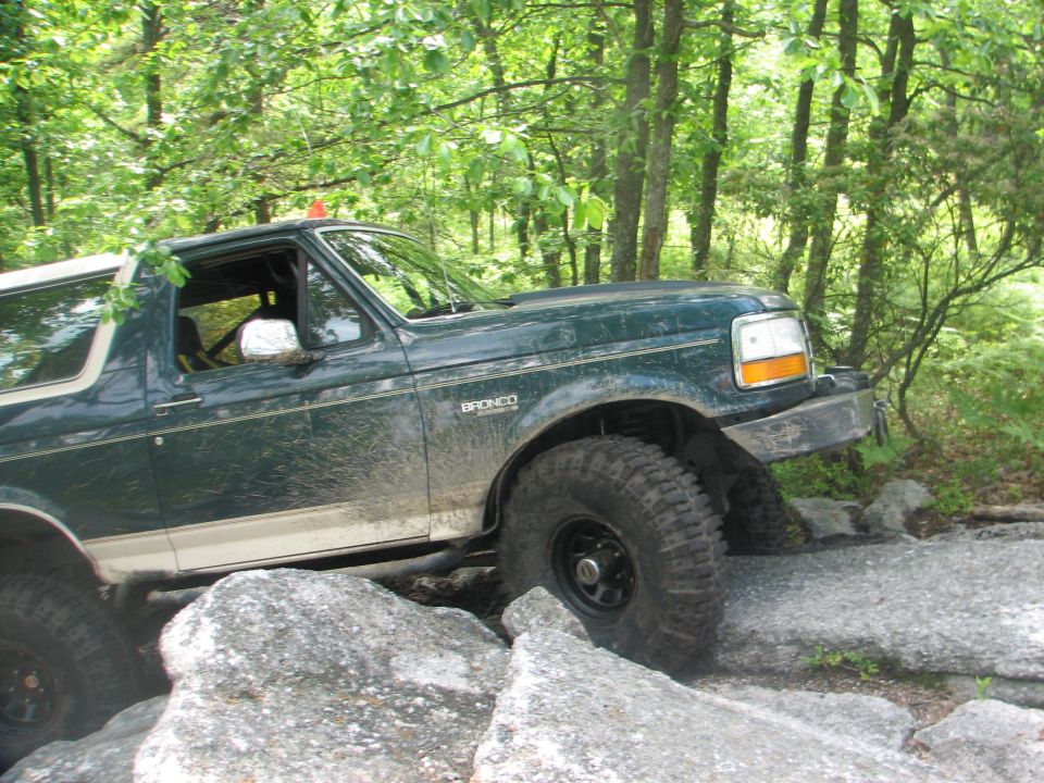 Muddy green and tan lifted Ford Bronco crawling over rock ledges on a wooded trail