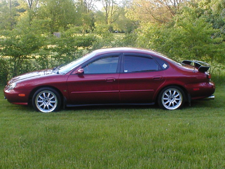 Dark red Ford sedan in profile on grass, rear spoiler, chrome wheels, green hedge behind
