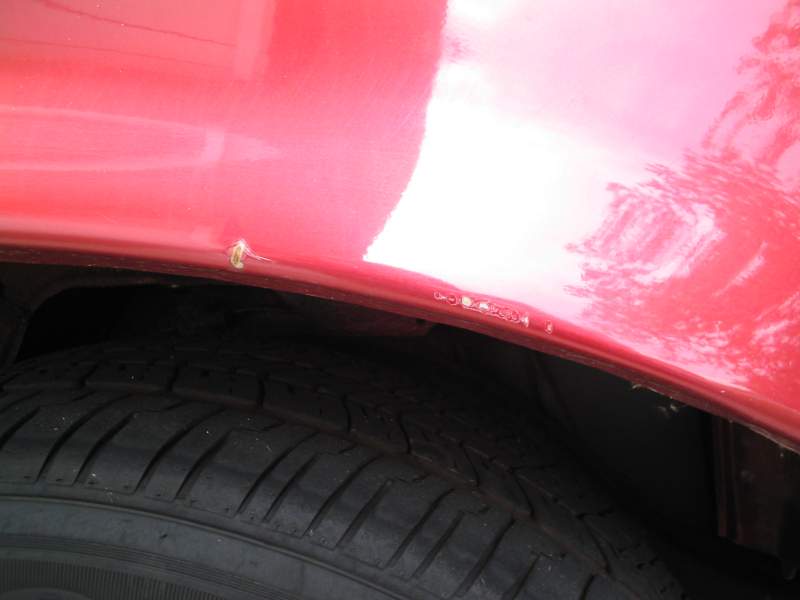 Close-up of red body panel edge above a tire showing chipped paint and a stud