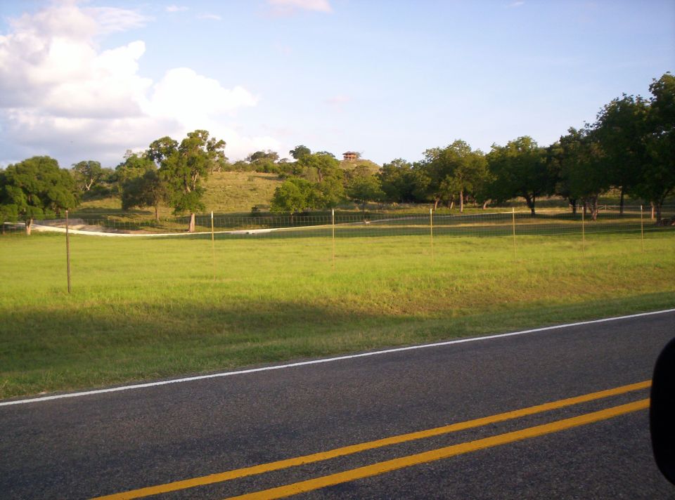 View of a grassy field and trees with a building on a distant hill, seen from a roadside