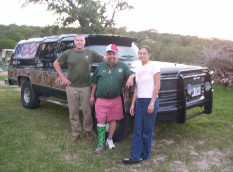 1986 Ford Bronco - Three people stand next to a customized black SUV with camouflage body panels and an American flag decal on the rear window.