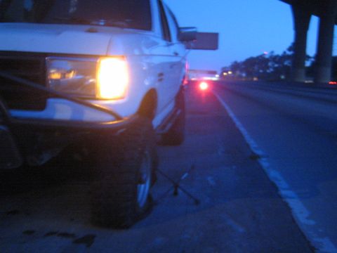 1990 Ford Bronco - white lifted truck on a highway shoulder at dusk with its headlight lit and a four-way lug wrench resting beside the front tire.