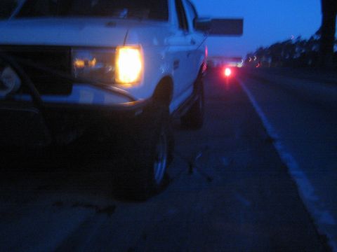 1990 Ford Bronco - white lifted truck stopped on a roadside at dusk, single headlight glowing, with distant traffic taillights along the highway.