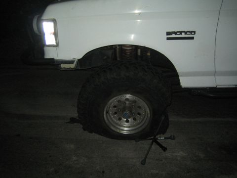 1990 Ford Bronco - Side view of a white lifted Ford Bronco's front wheel at night, with a large mud tire, exposed coil suspension, and a BRONCO fender badge.