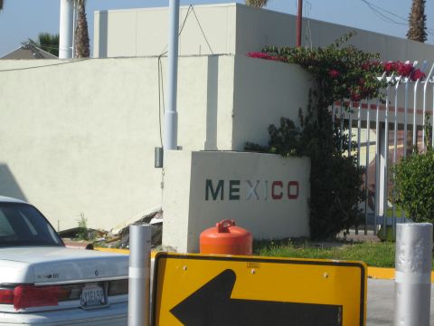 1990 Ford Bronco - roadside wall spelling MEXICO in the national colors stands behind a yellow left-arrow sign and a parked silver car.
